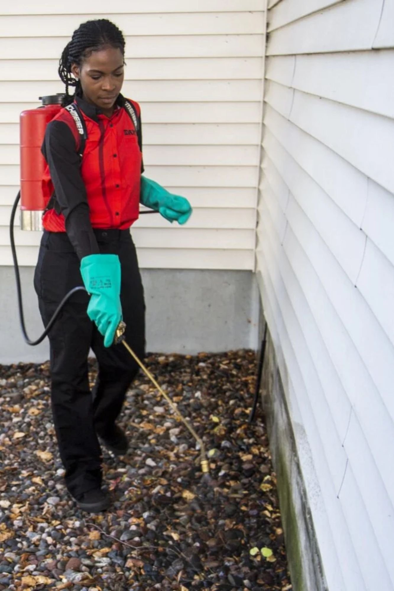 A woman in a red jacket and green gloves is spraying pesticide on a house during a pest control service in San Antonio, TX.